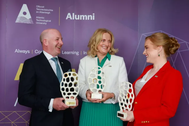 Three individuals standing in front of a purple backdrop with the Atlantic Technological University Alumni branding, each holding a trophy. They are dressed formally and smiling at each other.