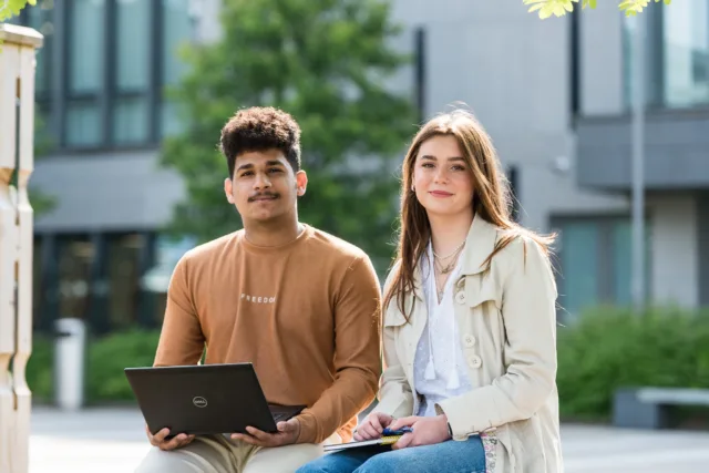 image of two students on a campus, one of the students is holding a laptop