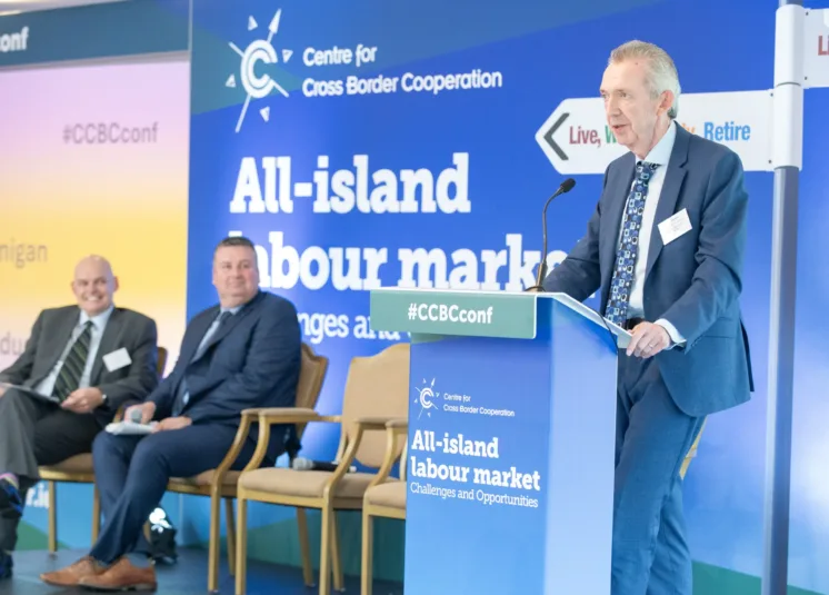 Paul Hannigan speaks at a cross border conference. Two other speakers sit on chair on nearby engaged in what he is saying.