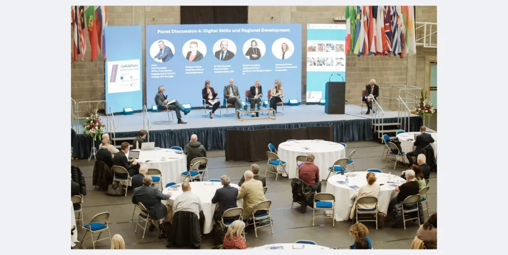 A panel discussion takes place on stage with an mc and four panellist sitting with a large digital screen behind them. In the audience there is circular tables with attendees.
