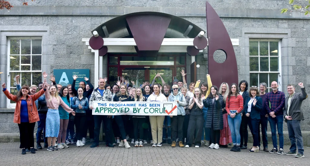 A group of people standing in front of a stone building with large windows and a distinctive maroon sculpture near the entrance. They are holding a banner that reads, “THIS PROGRAMME HAS BEEN APPROVED BY CORU” with a green checkmark.
