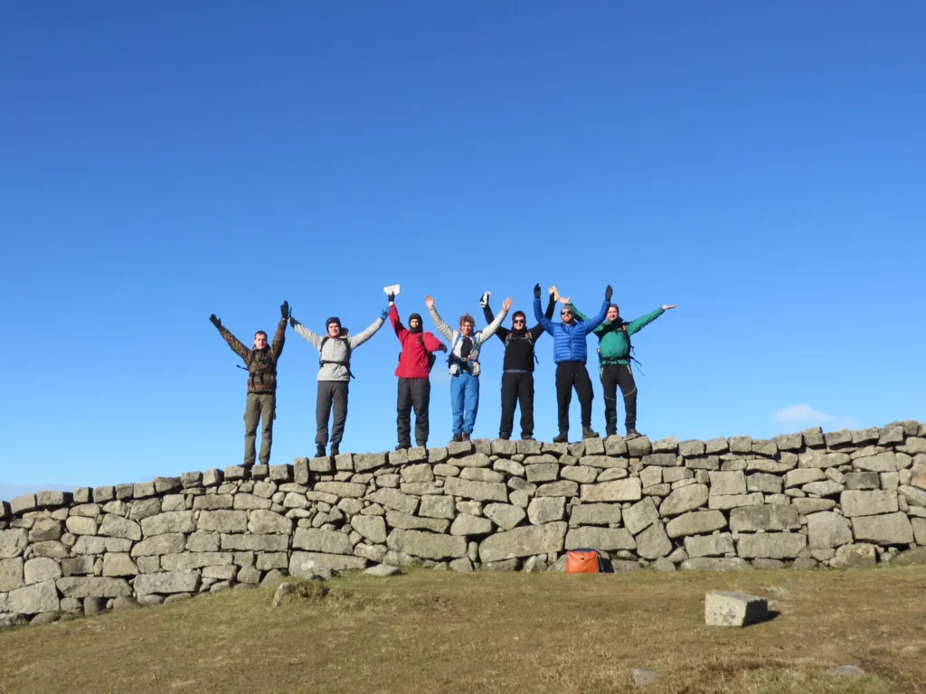 Six hikers standing on a stone wall with arms raised in celebration under a clear blue sky