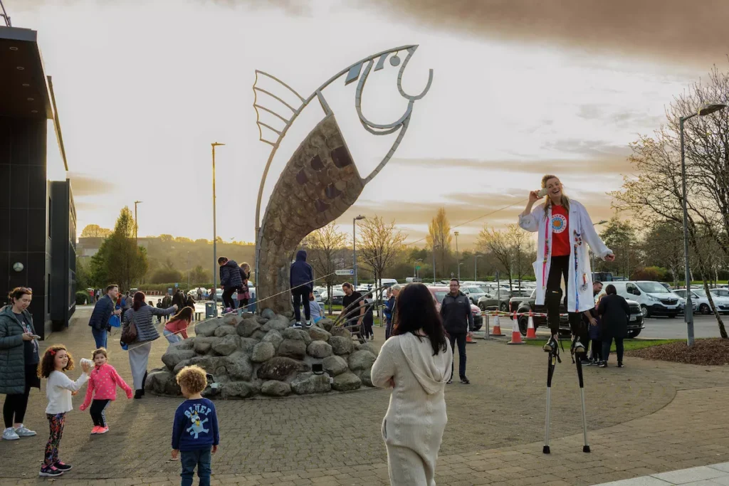 Outdoor scene with a large fish sculpture on rocks; people gathered around, and a performer on stilts entertaining the crowd.