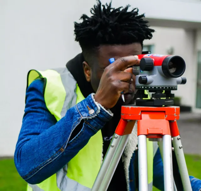 A person wearing a high-visibility safety vest and a denim jacket is operating a red and grey surveying instrument mounted on a tripod. The scene appears to be outdoors near a building with white walls.