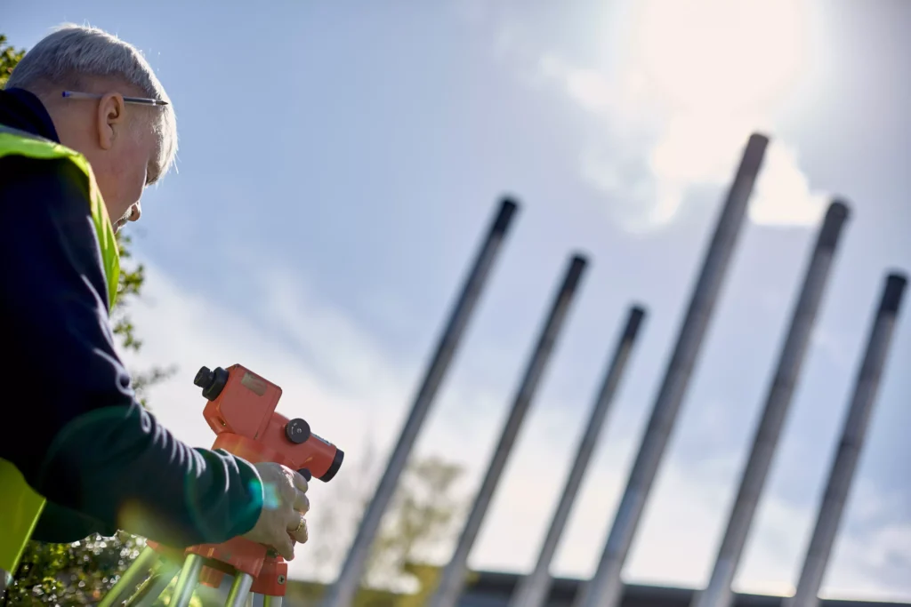 A person in a high-visibility vest is adjusting an orange surveying instrument on a tripod under bright sunlight. Several tall metal poles are visible in the background against a clear sky.