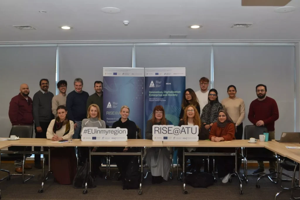 Large group of people gathered in a conference room, standing and sitting behind tables arranged in a U‑shape. Two vertical RISE@ATU programme banners stand at the back.