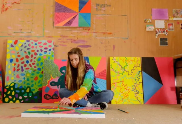 A person wearing a colorful tie-dye hoodie sits on the floor of an art studio, painting a bright abstract design on a canvas. Several large, vibrant abstract paintings lean against the wall behind them, featuring bold shapes, patterns, and vivid colors. The walls show paint marks and small artworks pinned up, creating a creative, expressive workspace.