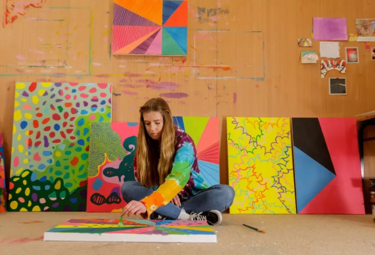 A person wearing a colorful tie-dye hoodie sits on the floor of an art studio, painting a bright abstract design on a canvas. Several large, vibrant abstract paintings lean against the wall behind them, featuring bold shapes, patterns, and vivid colors. The walls show paint marks and small artworks pinned up, creating a creative, expressive workspace.