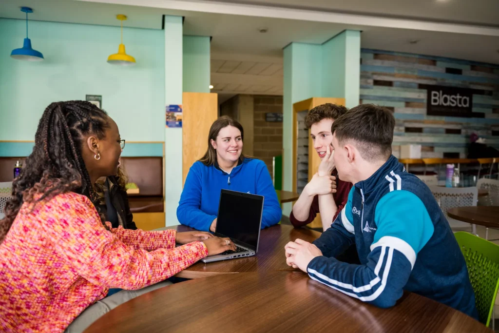 A group of four people sit around a wooden café table with an open laptop in front of them. The setting includes colourful hanging lights, cushioned seating, and a wall sign that reads “Blasta” in the background.