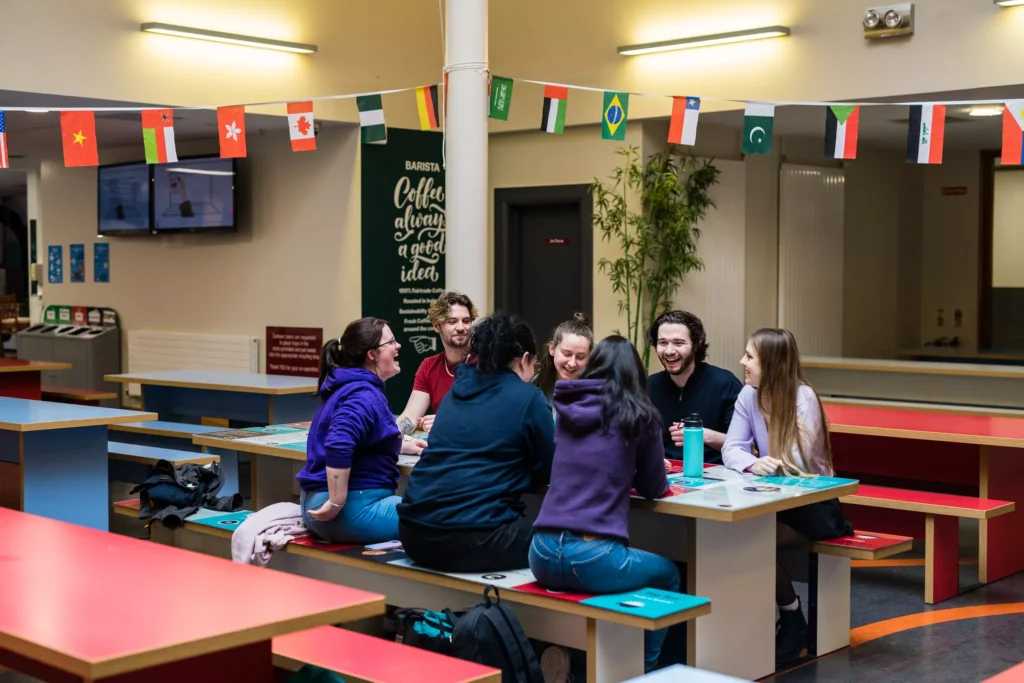 A group of students sit around a table in a campus café area, chatting and laughing beneath a string of international flags.