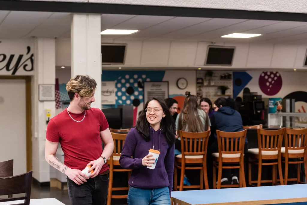 A student in a red t-shirt and a student in a purple hoodie are walking and smiling towards each other, holding cups in a cafeteria. Behind them, a group of people are seated at a table.
