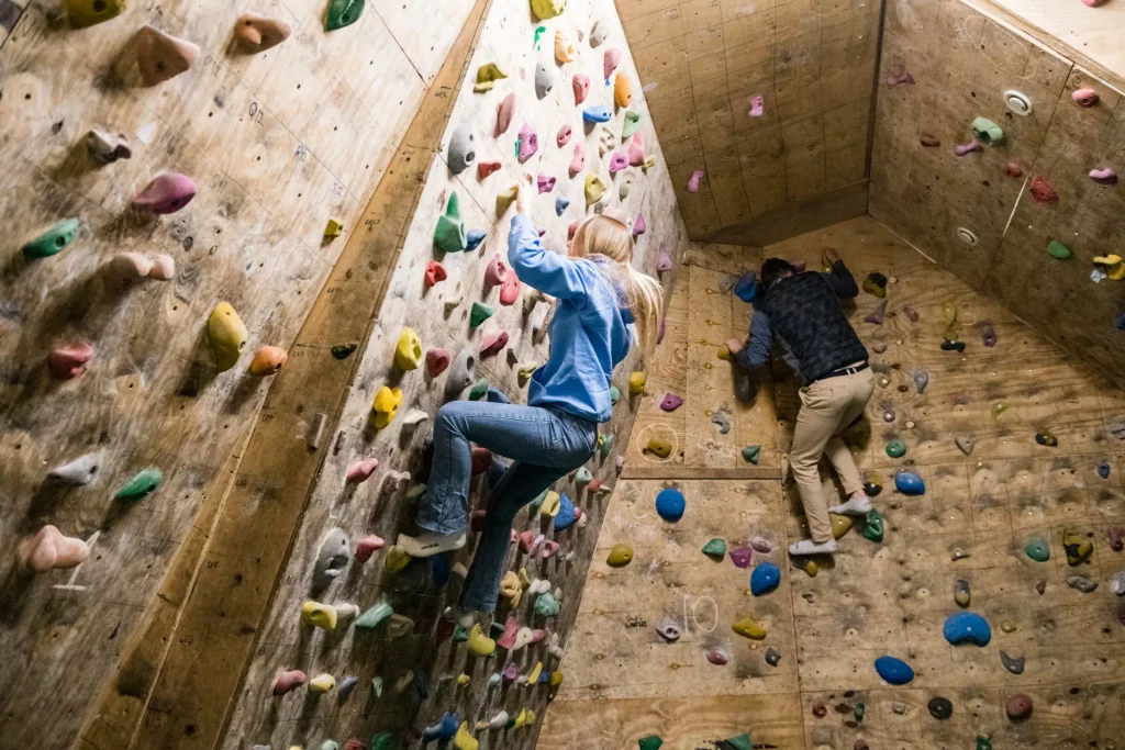 Two people climb a wooden indoor bouldering wall with colourful handholds.