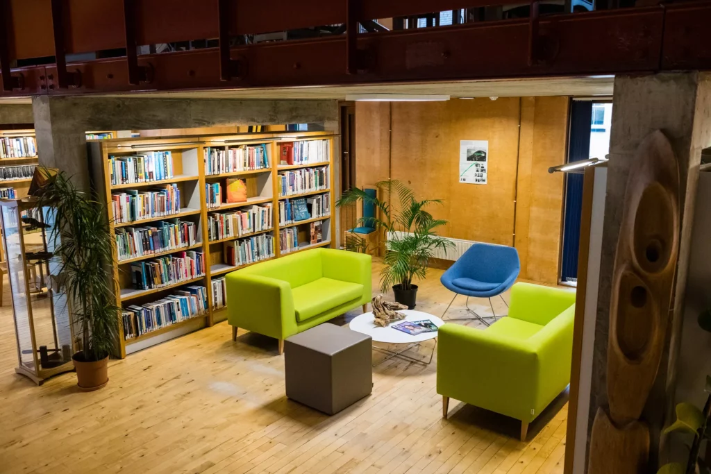 A cozy library seating area with wooden floors and bookshelves filled with books. Three brightly colored chairs—two green armchairs and one blue chair—surround a round white table. Several potted plants are placed around the space, and a wooden wall with a poster is visible in the background.