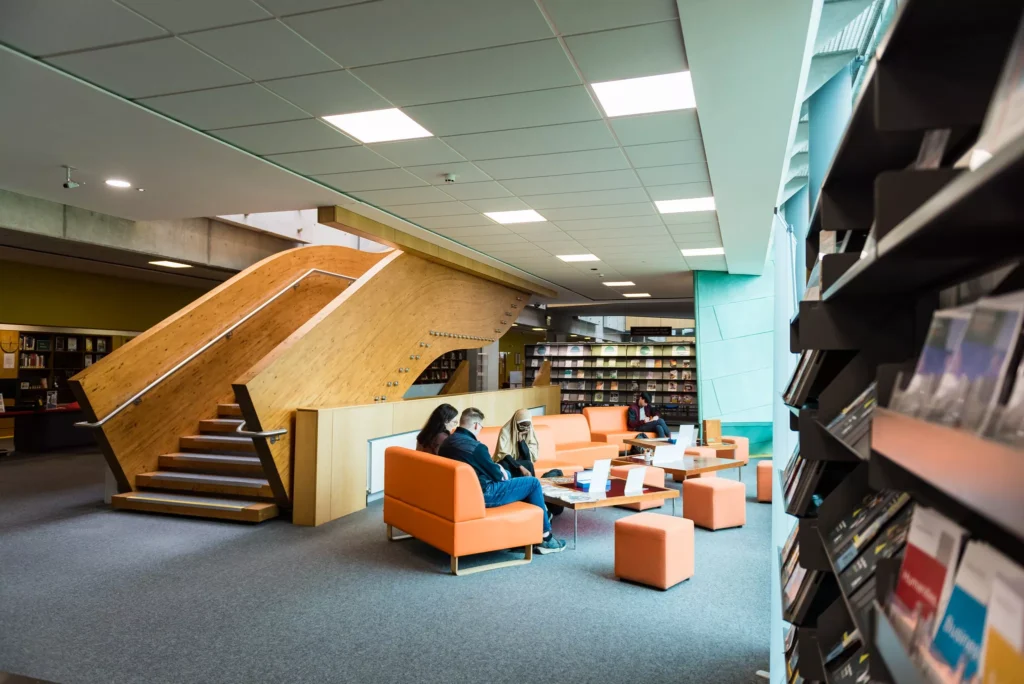 A modern library interior with wooden stairs and orange seating, where several people sit and read or study near low tables, surrounded by bookshelves and magazine racks.