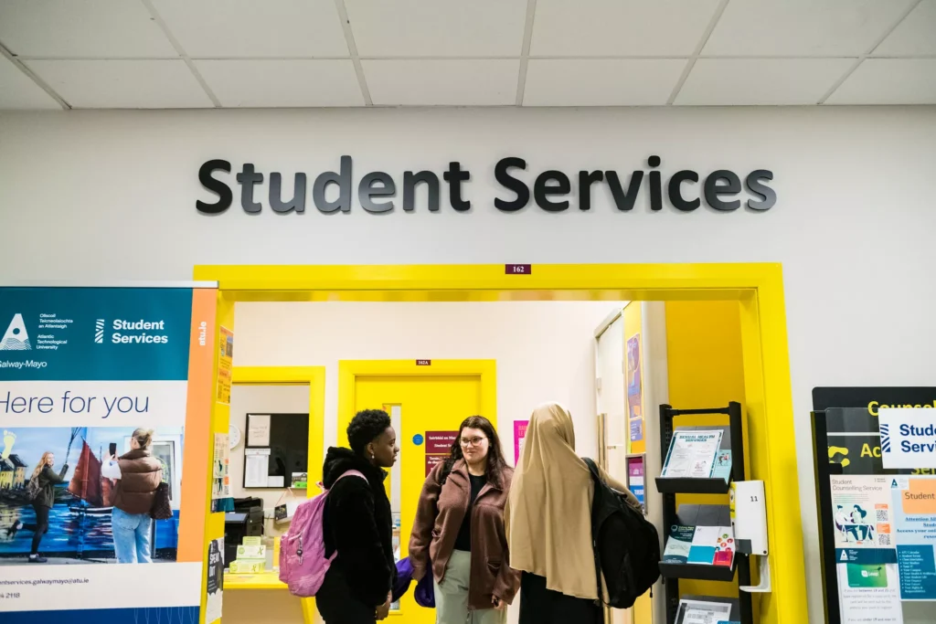 Three students stand talking at the entrance to a student services office with bright yellow door frames and a large “Student Services” sign above.