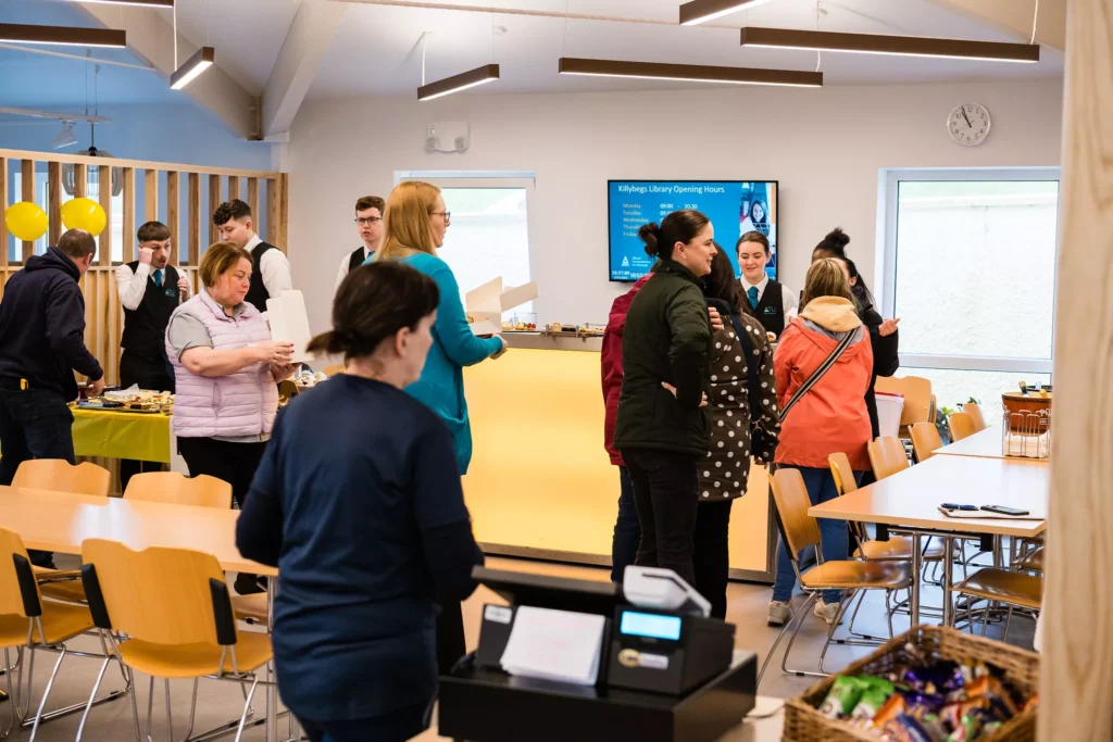 A busy café with several people standing near a long counter where food is displayed. Wooden chairs and tables fill the space, and a digital screen on the wall shows information about library opening hours. Overhead lights and natural light from windows brighten the room.