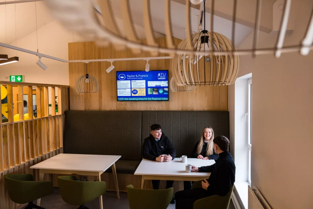 A small café-style seating area with three people sitting at a cushioned booth bench with white tables. A digital screen displaying announcements is mounted on a wooden slat wall behind them. Overhead, large decorative pendant lights hang from the ceiling, and the room features green chairs and warm neutral tones.