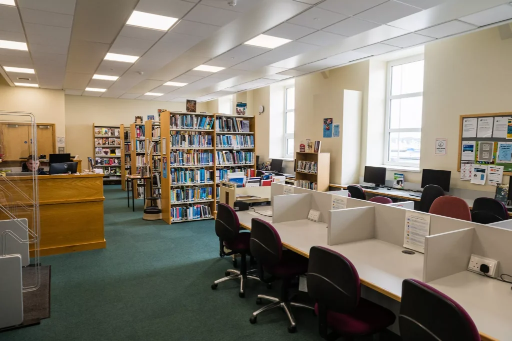 A bright library space with study desks, desktop computers, bookshelves, and a service desk. The room has high windows, overhead lighting, and green carpeted floors. Several shelves are filled with books, and noticeboards with posters and information are displayed on the walls.