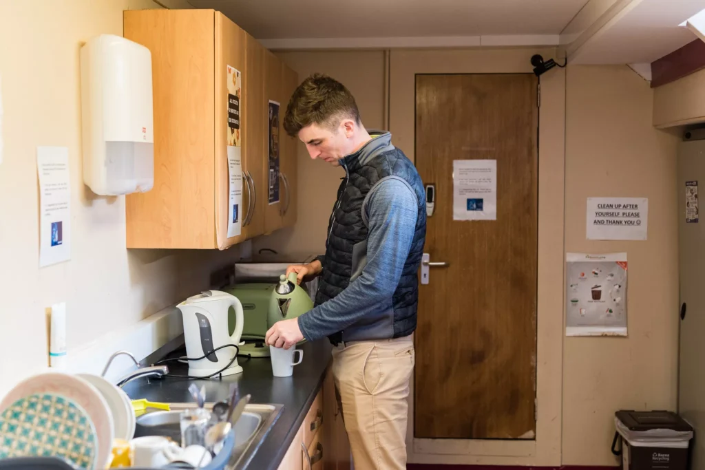A person stands in a kitchenette, pouring hot water from a kettle into a mug on the counter. The room has wooden cabinets, a sink with dishes, a microwave, and signs posted on the walls and door.
