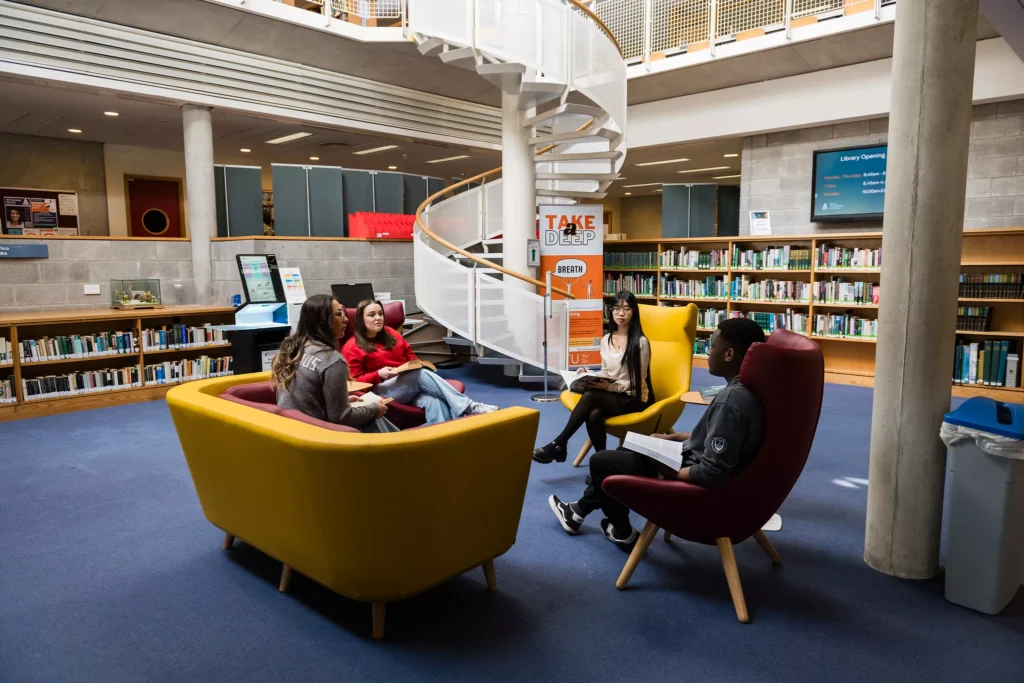 A group of four people sit in a circle on colourful lounge chairs in a library reading area. The space includes tall bookshelves, a blue carpet, and a white spiral staircase leading to an upper level.