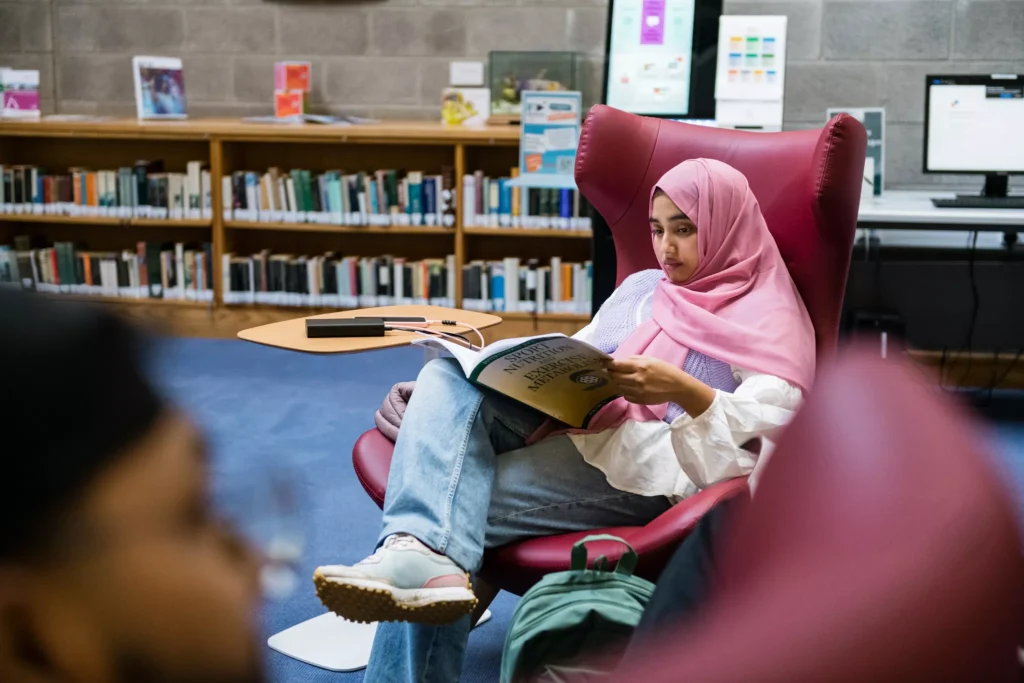 A person sits in a red lounge chair in a library, reading a book with crossed legs. Bookshelves, computers, and a table with devices are visible in the background. A backpack rests on the floor beside the chair.