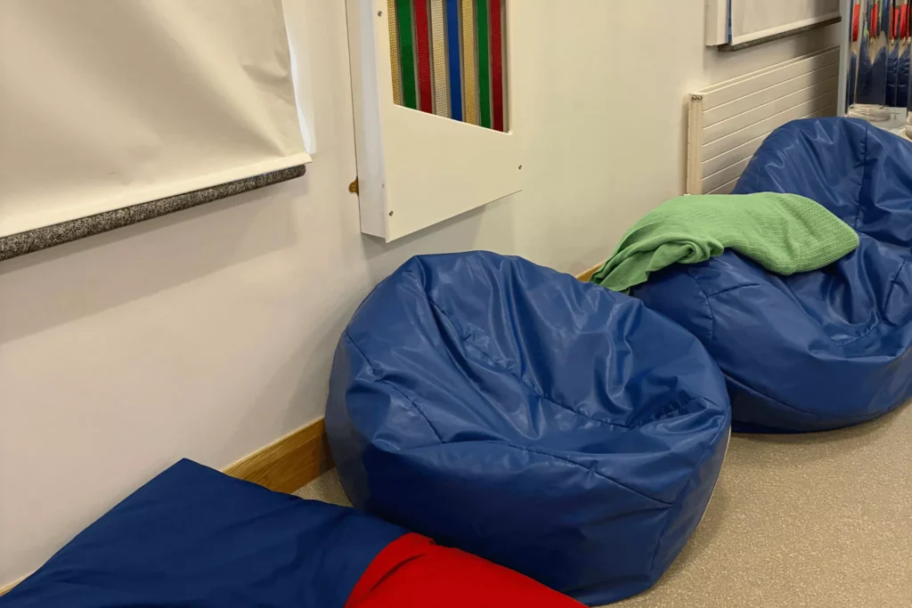 Several blue bean bags are placed in a room with a green towel resting on top of one. There is also a multi-coloured striped mesh panel on the wall.