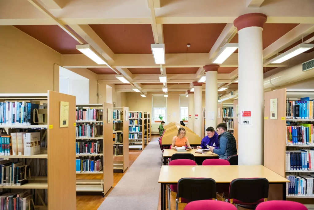 A quiet library with rows of bookshelves and large white columns. Three people sit together at a table in the centre, looking at books or notes, while additional shelves and study tables fill the background.