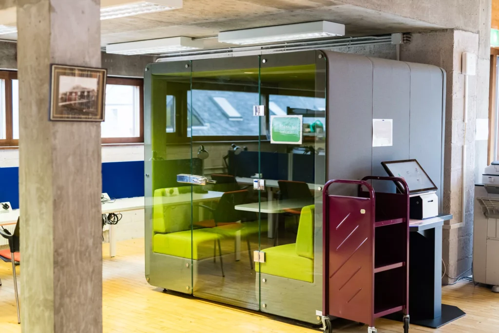 A small modern study pod with glass walls sits in an open library or workspace. Inside the pod are a table and bright green chairs.