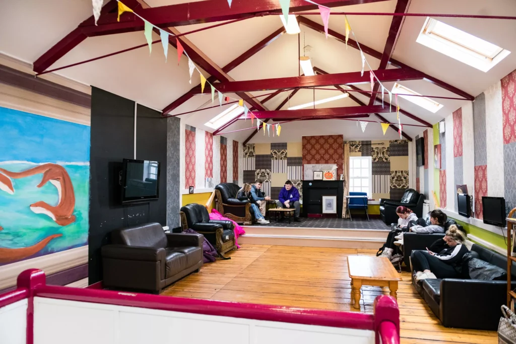 A spacious social room with wooden floors, leather sofas, and colorful bunting hanging from exposed ceiling beams. Several people sit on the sofas around the room, while a small group gathers at a table in the back. A large mural and a wall-mounted TV are visible on the left side.