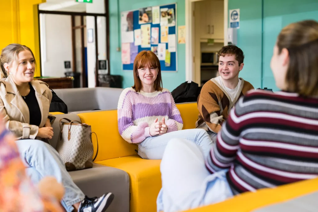 A group of people sit together on yellow and grey couches in a bright social space. Colourful walls, a noticeboard with posters, and a doorway to an adjoining room are visible in the background.
