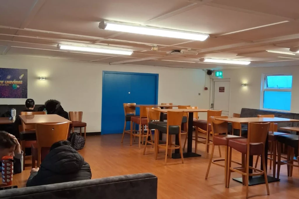 A room with tall wooden tables and chairs, some of which are occupied by people. There is a blue door on the right side of the room and a colourful poster on the wall.
