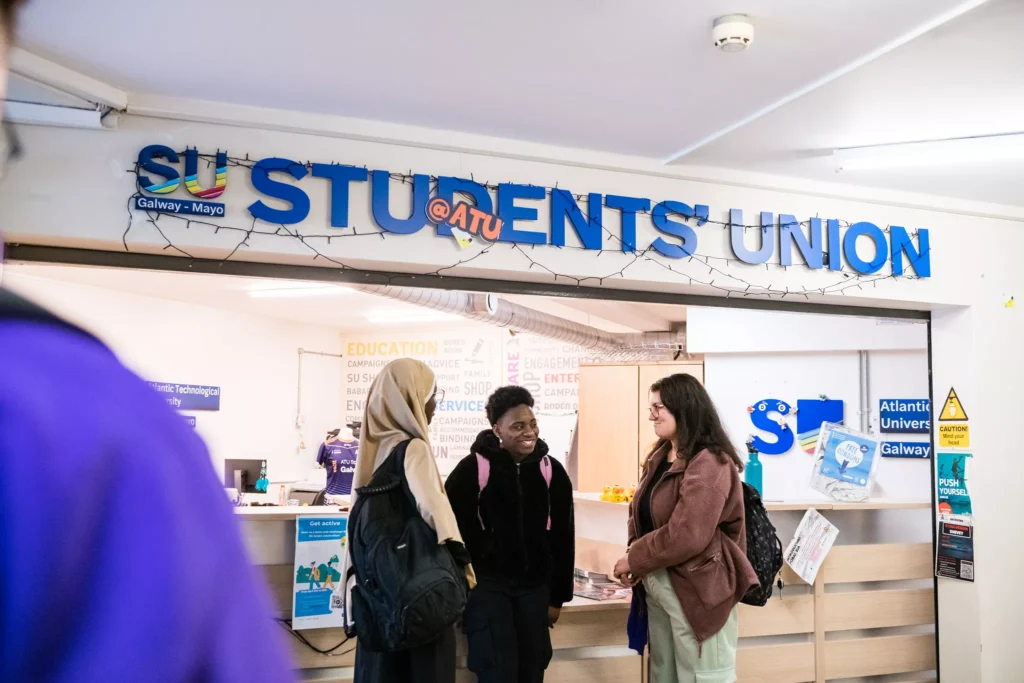 Three students with backpacks stand chatting and smiling at the reception area beneath a large blue “Students’ Union” sign.