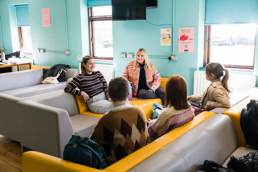 A group of five people sit together on yellow and grey sofas in a bright lounge area with turquoise walls. The room includes windows with blue blinds, wall posters, a mounted TV, and a wooden floor.