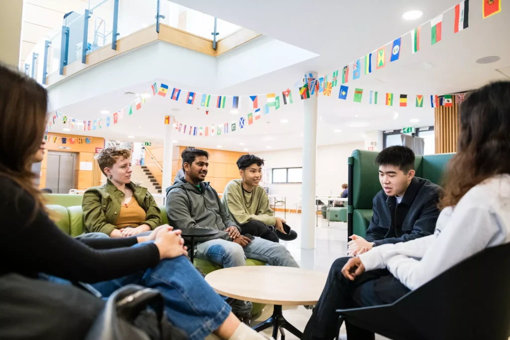 A group of students sit in a circle in a bright university lounge, talking together beneath rows of international flags hanging across the atrium.