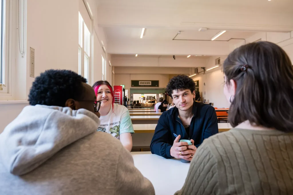 A group of four people sit around a white table in a bright cafeteria, with long tables and a serving area visible in the background.