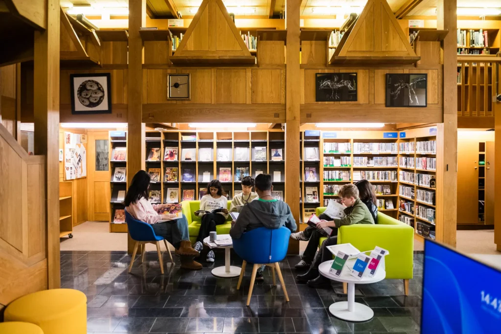 A brightly lit library space with wooden walls and bookshelves filled with books and magazines. In the center, six people sit in a circle on colorful chairs—blue, green, and lime—reading or looking at materials. Small round tables hold books and pamphlets. Artwork is mounted on the upper wooden structure, and additional shelves line the background.