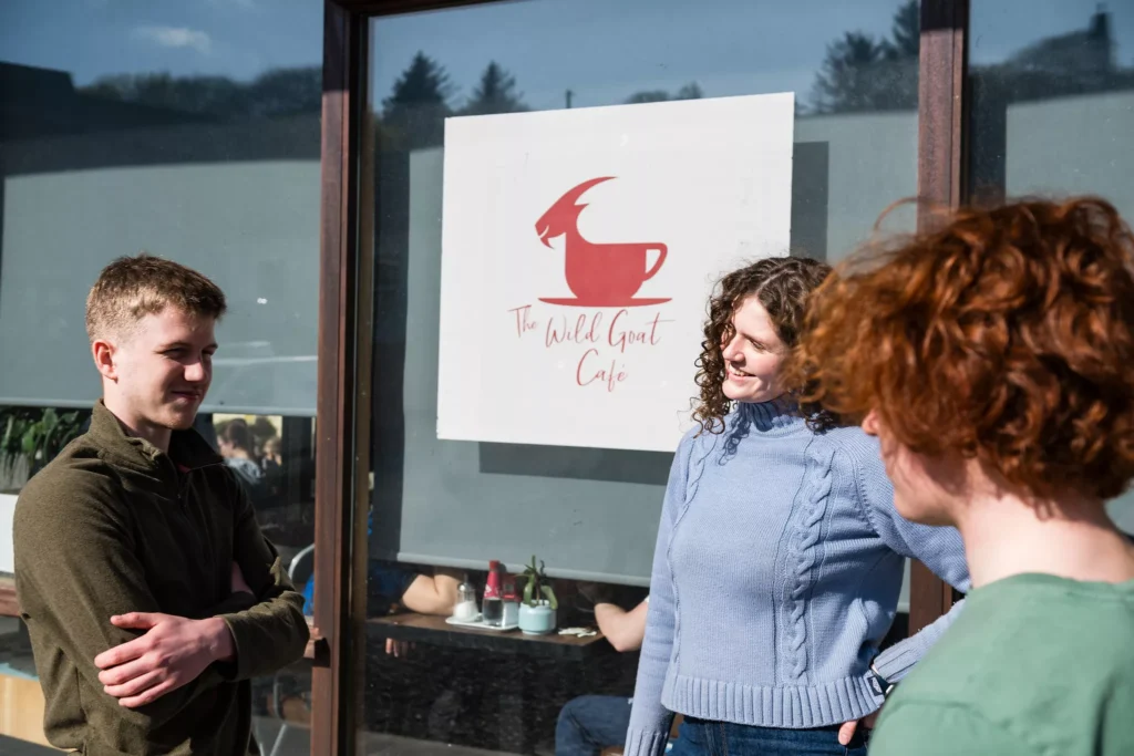 A group of three people stand outside a café window featuring a sign with a red goat-shaped coffee cup and the text “The Wild Goat Café.”