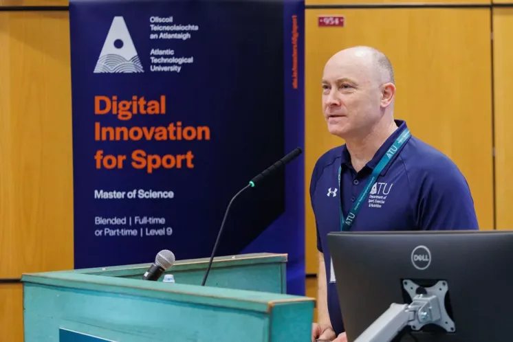 A person standing at a lectern with a microphone and a computer monitor, speaking in front of a banner reading “Digital Innovation for Sport” and “Atlantic Technological University,”.