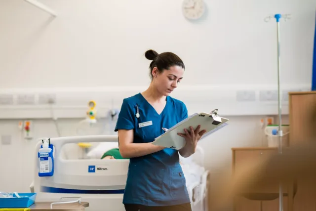 A person in blue scrubs stands beside a hospital bed, writing on a clipboard. Medical equipment, wall outlets, and a clock are visible in the clinical room.
