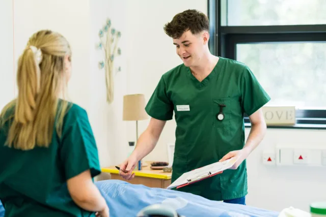 Two people in green scrubs stand beside a treatment bed in a clinical room, with one person holding a clipboard and pen. A window, wall fixtures, and small furnishings are visible in the background.