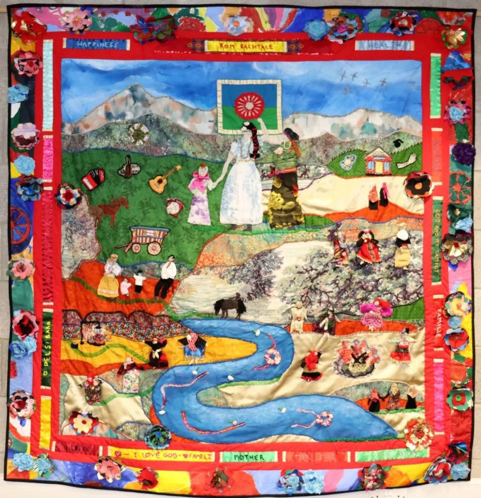 A colourful textile tapestry showing multiple small scenes across a landscape with hills, fields, a winding river, animals, buildings, musical instruments and groups of people engaged in everyday activities, all bordered by a patterned frame with fabric flowers.