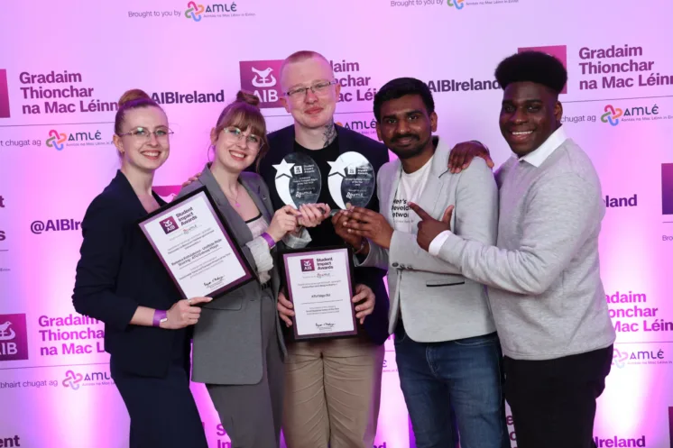 Photograph of five people standing together in front of a backdrop with "Gradaim Thionchar na Mac Léinn" and "AIB Ireland" logos, holding awards and certificates. The group are celebrating an achievement, with two individuals holding plaques and others smiling and posing for the photo.