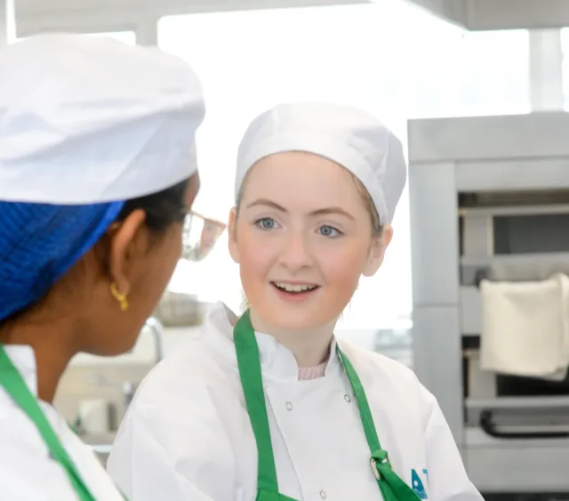 Photograph of two teens wearing white chef uniforms, green aprons, and white hats in a commercial kitchen setting. One chef is facing the camera with a blurred face, while the other is turned sideways, suggesting a conversation or collaboration during food preparation.