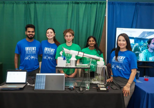 Photograph of five people standing behind a table displaying a science project with laptops, a mechanical model, and electronic components. Participants wear blue and green shirts with "Invent for the Planet".