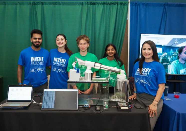 Photograph of five people standing behind a table displaying a science project with laptops, a mechanical model, and electronic components. Participants wear blue and green shirts with "Invent for the Planet".