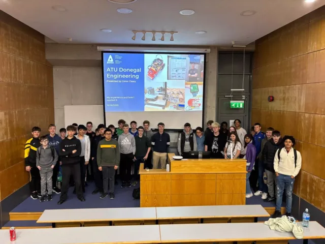 A large group of people stand together at the front of a lecture theatre behind a wooden lectern, with a projection screen and tiered seating visible.