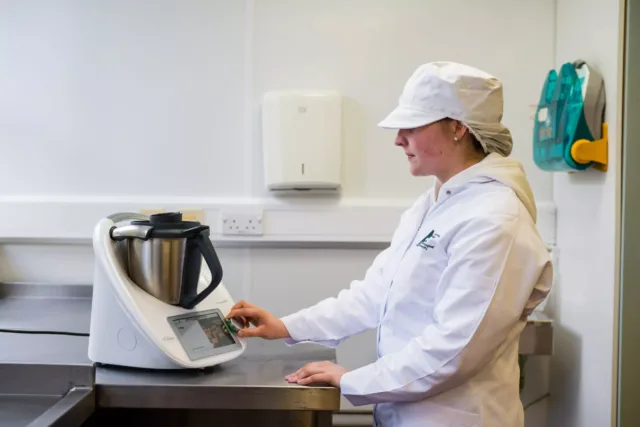A person wearing protective clothing stands at a stainless steel counter, operating a countertop food‑processing appliance in a clean, white room.
