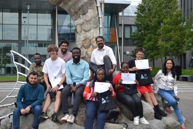 A group of people seated on rocks outdoors, holding certificates and black tote bags, with a modern glass building and a large metal sculpture in the background.