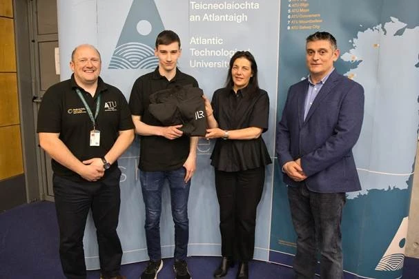 Photograph of four people standing indoors in front of a backdrop featuring Atlantic Technological University branding and a map.