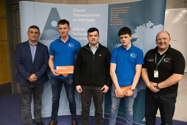 Photograph of five men standing in front of a blue backdrop displaying Atlantic Technological University logo and text in English and Irish. Two students wear blue ATU polo shirts holding orange boxes